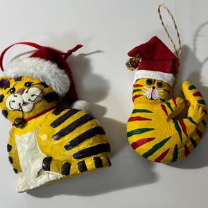 May include: Two Christmas cat ornaments. One is a yellow cat with black stripes, wearing a red Santa hat with white trim and a gold bell. The other is a curled-up yellow cat with red and green stripes, also wearing a Santa hat.