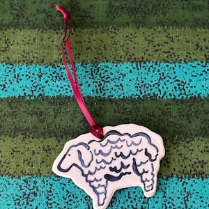 Könnte beinhalten: Ein weißes Keramik-Schaf-Ornament mit dunkelblauen Umrissen und Details. Das Schaf hat ein rosa Band zum Aufhängen. Das Ornament befindet sich auf einem Hintergrund aus türkis, grünen und schwarzen Streifen.