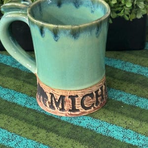 May include: A ceramic mug with a glossy turquoise and blue glaze on the upper portion and a textured, light brown base. The lower section features the black embossed text MICH, partially visible, along with a small black silhouette of an animal.
