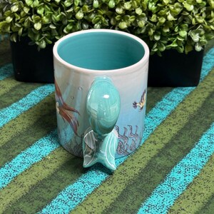 May include: A ceramic mug with a light blue exterior featuring an underwater scene with seaweed and a fish. The interior is a solid turquoise color. The handle is shaped like a mermaid tail, colored in a glossy turquoise with white line details.