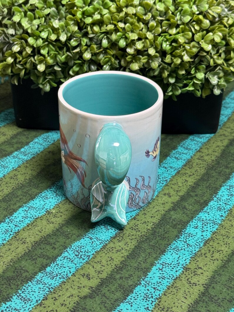 May include: A ceramic mug with a light blue exterior featuring an underwater scene with seaweed and a fish. The interior is a solid turquoise color. The handle is shaped like a mermaid tail, colored in a glossy turquoise with white line details.