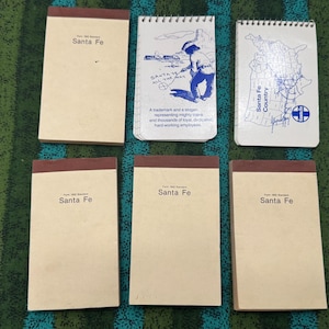 Pode incluir: Seis blocos de notas retangulares com cabeçalhos castanhos. Quatro blocos são de cor creme com o texto Santa Fe. Um bloco apresenta uma ilustração azul de uma pessoa e o texto Santa Fe All The Way. Um bloco mostra um mapa azul Santa Fe Country.