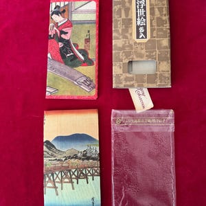 May include: Four rectangular items on a red surface. Two are printed with Japanese art, one featuring a woman playing an instrument and the other a bridge over water. A box with Japanese text and a clear plastic sleeve are also present.