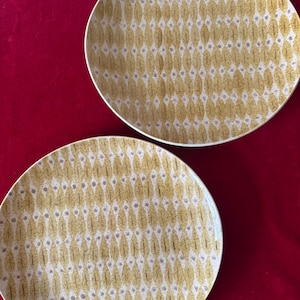 May include: Two yellow and white ceramic dinner plates with a geometric pattern. The plates have a white background with a pattern of yellow and brown leaves.