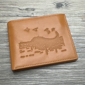 May include: A tan leather wallet with a detailed embossed design featuring a landscape with birds and trees. The wallet is rectangular and has a stitched border.