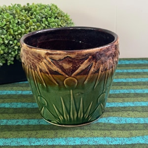 May include: A ceramic planter featuring a two-tone glaze with a dark brown upper section and a vibrant green lower section. The exterior surface is decorated with a raised, embossed sunburst pattern consisting of radiating lines and circles.