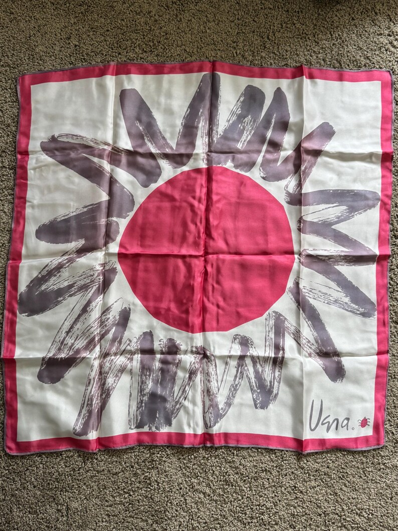 May include: Square silk scarf with a sunburst design. The scarf features a large pink circle in the center, surrounded by gray brushstroke-style rays on a white background. A pink border frames the design. The scarf has the word "Usna" in the corner.