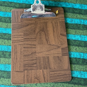 May include: A rectangular clipboard with a brown wood grain pattern. The metal clip is silver and has the letter "A" on it. The clipboard is on a green and blue striped surface.
