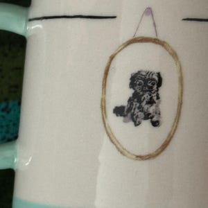 May include: White ceramic mug with a light teal handle and base. The mug features a framed illustration of a small, black and white dog. A thin black line is above the framed dog illustration.