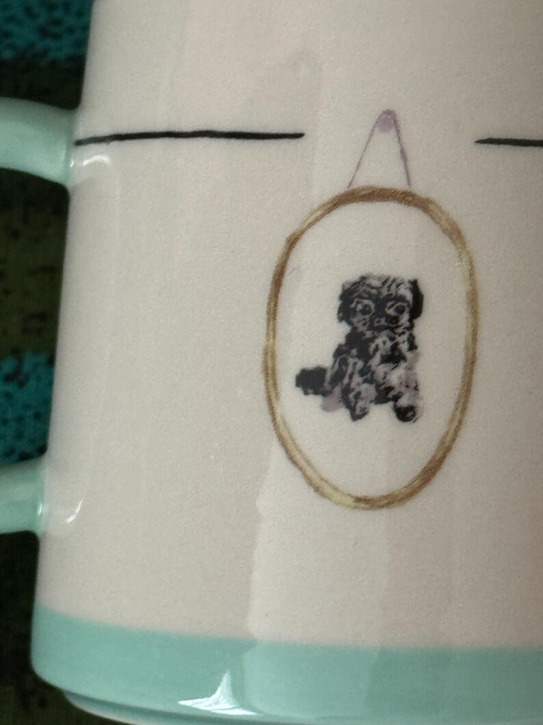 May include: White ceramic mug with a light teal handle and base. The mug features a framed illustration of a small, black and white dog. A thin black line is above the framed dog illustration.