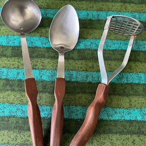May include: Three vintage kitchen utensils: a ladle, a spoon, and a potato masher. Each utensil has a stainless steel head and a dark brown wooden handle. The items are arranged on a green and blue striped surface.