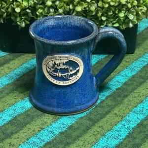 May include: A cobalt blue ceramic mug with a glossy, mottled finish and a curved handle. An oval-shaped white medallion is centered on the front, featuring a black line drawing of cave formations and the text Blanchard Springs Caverns Fifty-Six Arkansas.