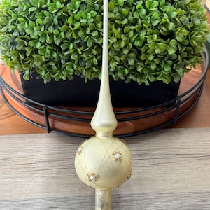 May include: A vintage-style Christmas tree topper. The topper is primarily cream-colored with gold accents, including a decorative gold chain and small pearl-like embellishments. The topper has a long, slender spire and a round base.