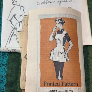 May include: A vintage sewing pattern envelope for a cobbler apron, featuring a black and white illustration of a person wearing the apron over a dark outfit. The envelope includes the text Printed Pattern, 4853, and Pattern Department address.