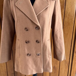 May include: A tan double-breasted coat with a notched collar and six brown buttons. The coat has two front pockets and long sleeves. The coat is displayed on a black mannequin.