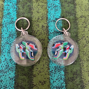 May include: Two clear acrylic keychains with a colorful sneaker design. Each keychain features a silver keyring and a circular pendant with a graphic of sneakers in red, green, blue, and white.