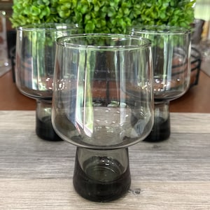 May include: Four clear glass tumblers with a dark gray base and stem. The glasses have a rounded bowl shape and are arranged on a wooden surface. A green plant is visible in the background.