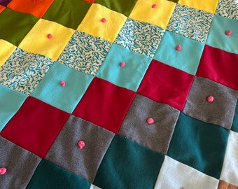 May include: A patchwork quilt with a variety of colors and patterns, including red, green, blue, yellow, and brown. The quilt is made of different fabrics and has pink pom-poms sewn on each square.