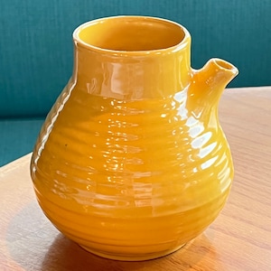 May include: A yellow ceramic pitcher with a rounded body and a small spout. The pitcher has a textured surface and a glossy finish.