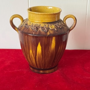 May include: A brown ceramic vase with two handles and a yellow rim. The vase has a decorative floral band around the top and a glossy finish. The vase is sitting on a red surface.