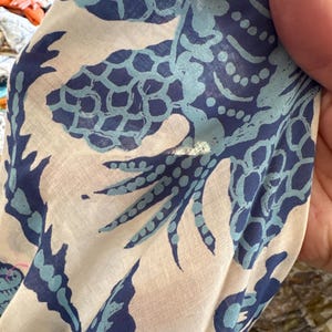 May include: Close-up of a white fabric with a repeating pattern of blue and light blue stylized sea creatures. The fabric is slightly wrinkled, and the design appears to be printed on the material. The fabric is being held by a person.