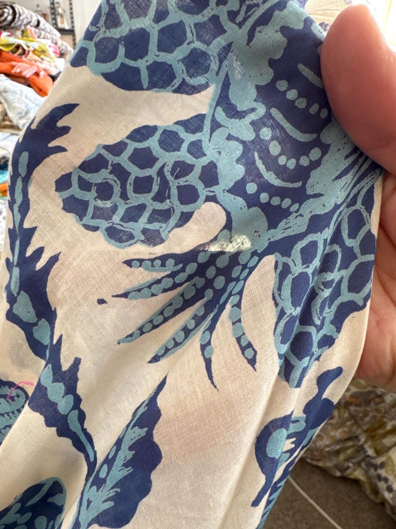 May include: Close-up of a white fabric with a repeating pattern of blue and light blue stylized sea creatures. The fabric is slightly wrinkled, and the design appears to be printed on the material. The fabric is being held by a person.