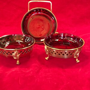 May include: Three small, round, ruby red glass dishes with gold metal bases and feet. The dishes are on a red velvet surface.