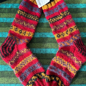 Op de afbeelding: Een paar kleurrijke gebreide sokken met een aangehecht label. De sokken hebben horizontale strepen in rood, geel, paars en zwart. Het label leest "newari studio". Deze warme sokken zijn perfect voor koud weer.