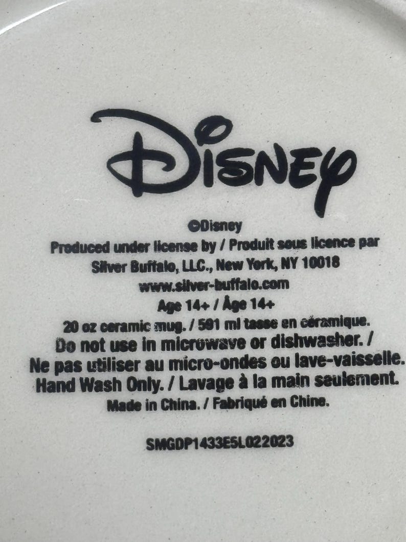 May include: A white ceramic mug base featuring black printed text that reads Disney, Produced under license by Silver Buffalo, LLC., New York, NY 10018, 20 oz ceramic mug, Do not use in microwave or dishwasher, Hand Wash Only, Made in China, SMGDP1433E5L022023.