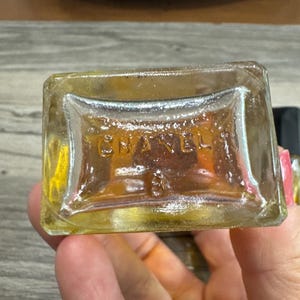 May include: A clear, rectangular perfume bottle with the embossed word "CHANEL" and the number "3" visible on the bottom. The bottle contains a golden-colored liquid. The image is a close-up shot.