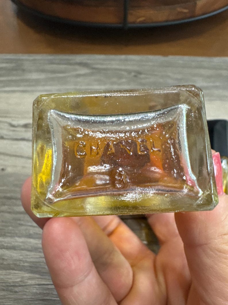 May include: A clear, rectangular perfume bottle with the embossed word "CHANEL" and the number "3" visible on the bottom. The bottle contains a golden-colored liquid. The image is a close-up shot.