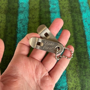 May include: A small, silver-colored "Quad Fold" tool with a key chain. The tool is held in a hand against a green and blue striped background. The tool has a hinged design and the words "Quad Fold" are engraved on it.