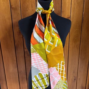 May include: A colorful silk scarf with a geometric pattern in shades of orange, yellow, pink, and green. The scarf is draped over a black mannequin, showcasing its design. The scarf's design includes stripes, squares, and other shapes.