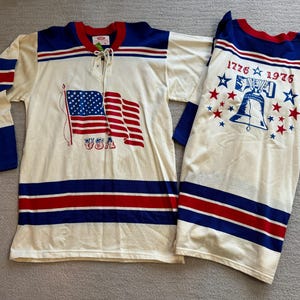May include: A white hockey jersey with red and blue stripes. The front of the jersey features a graphic of the American flag with the text "USA". The back of the jersey has the text "1776 1976" with a graphic of the Liberty Bell and stars.