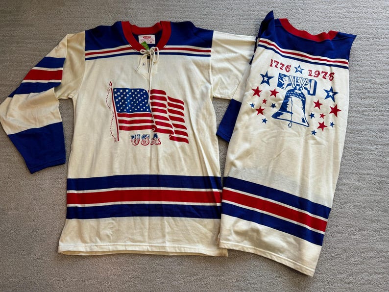 May include: A white hockey jersey with red and blue stripes. The front of the jersey features a graphic of the American flag with the text "USA". The back of the jersey has the text "1776 1976" with a graphic of the Liberty Bell and stars.