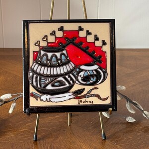 May include: A ceramic tile with a black frame depicting two Native American pottery vessels with geometric designs in black, white, blue, and red. The tile is on a gold metal easel and signed "Muhna".
