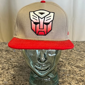 May include: A gray and red baseball cap with a white and red embroidered Transformers logo on the front.