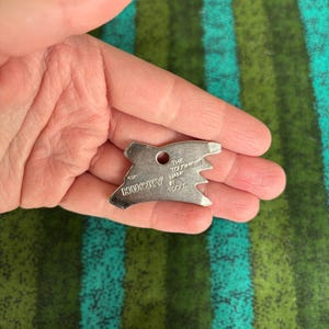 May include: A small, silver-colored Husky multi-tool with a hole and a notched edge. The tool has the words "HUSKY" and "THE TOUGHEST NAME IN TOOLS" stamped on it. The tool is held in a hand against a green and blue striped background.