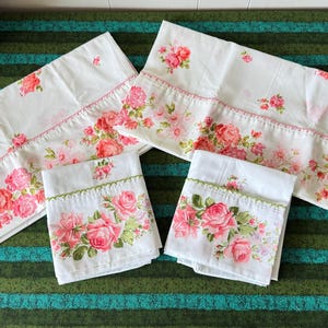 May include: White bedding set with a floral rose pattern in shades of pink and green. The set includes two pillowcases and two sheets, with decorative stitching along the edges. The bedding is arranged on a green and teal striped surface.