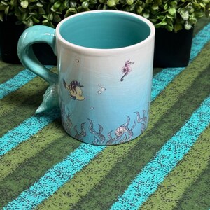May include: A ceramic mug with a light blue and white gradient exterior featuring illustrations of a small fish, a seahorse, and seaweed. The interior of the mug is a solid turquoise color, and the handle is shaped like a curved mermaid tail.