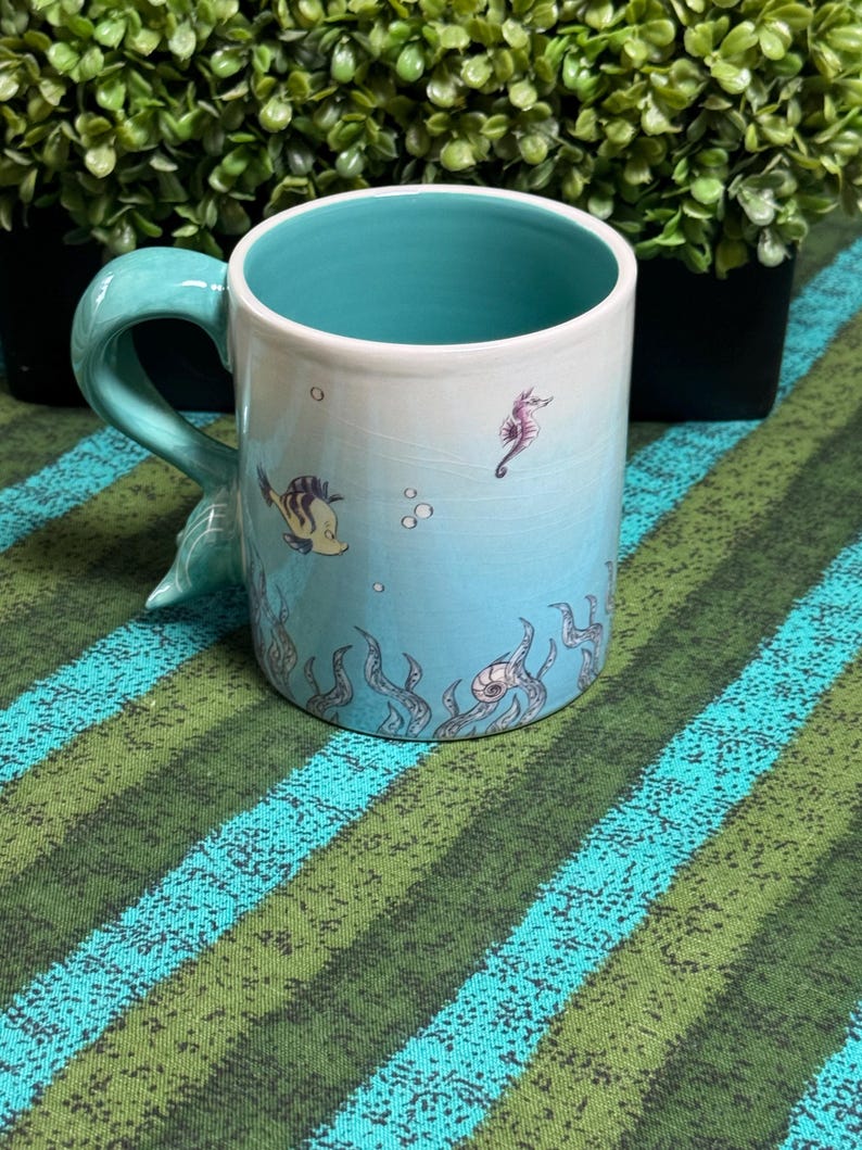 May include: A ceramic mug with a light blue and white gradient exterior featuring illustrations of a small fish, a seahorse, and seaweed. The interior of the mug is a solid turquoise color, and the handle is shaped like a curved mermaid tail.