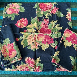 May include: A dark blue floral tablecloth with matching napkins. The fabric features a pattern of pink and red flowers, green leaves, and yellow grapes. The tablecloth and napkins are arranged on a striped blue and green surface.