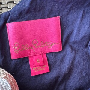 Pu&ograve; includere: Primo piano di un'etichetta rosa con testo dorato "Lilly Pulitzer" e un'etichetta taglia 6. L'etichetta &egrave; attaccata a un tessuto blu navy con finiture dorate.