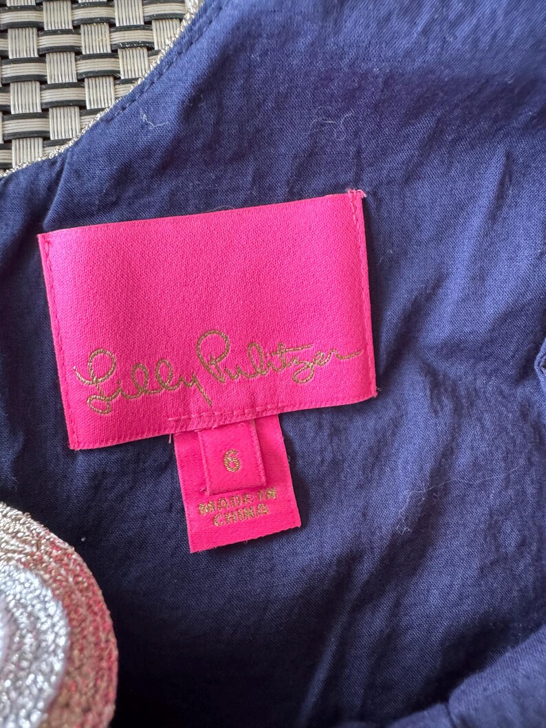 Pu&ograve; includere: Primo piano di un'etichetta rosa con testo dorato "Lilly Pulitzer" e un'etichetta taglia 6. L'etichetta &egrave; attaccata a un tessuto blu navy con finiture dorate.
