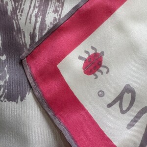 May include: Silk scarf with a white background, a red border, and a ladybug illustration. The scarf has a gray and white abstract design on one side. The scarf is folded, showing the corner with the ladybug.