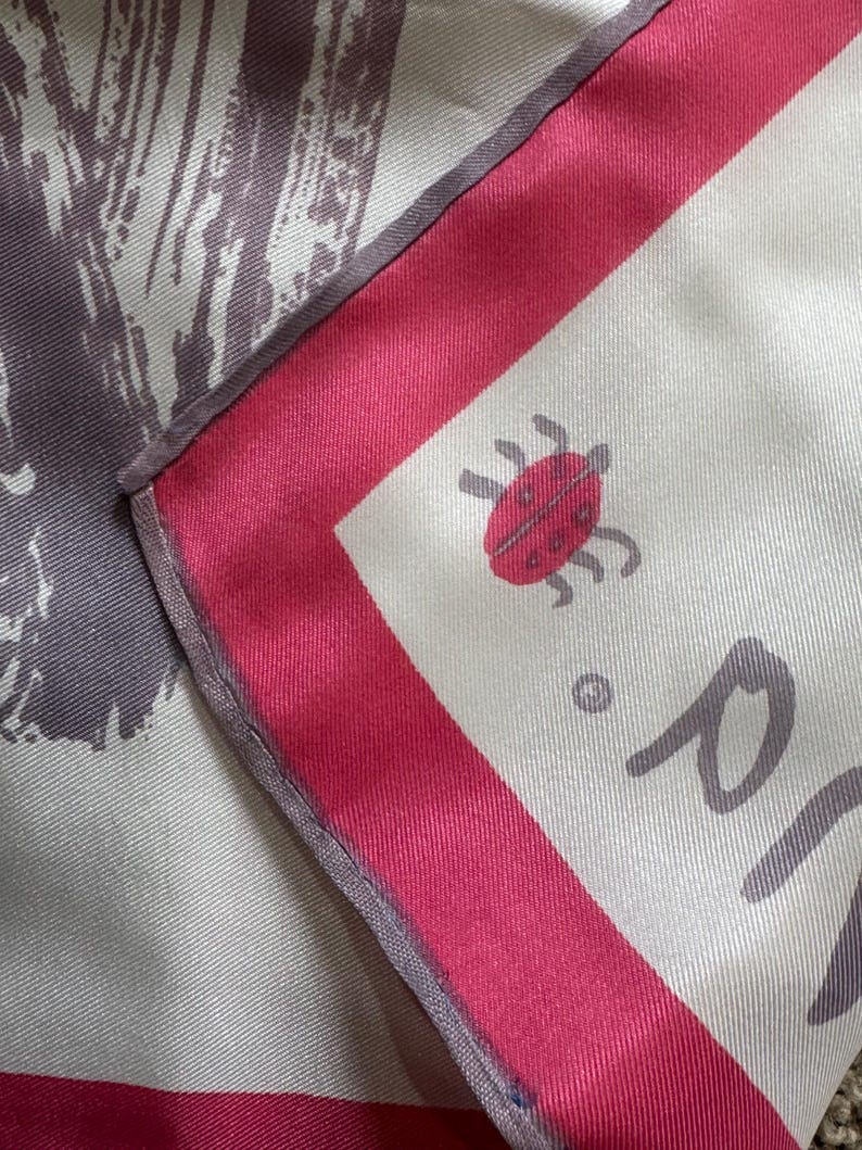 May include: Silk scarf with a white background, a red border, and a ladybug illustration. The scarf has a gray and white abstract design on one side. The scarf is folded, showing the corner with the ladybug.