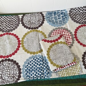 May include: A beige fabric with a repeating pattern of large, colourful circles. The circles feature various designs and colours, including red, blue, grey, and brown. A folded pillow with a matching design is also visible.