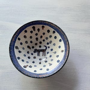 May include: A small white ceramic bowl with a blue rim and a pattern of blue dots. A black silhouette of a dog is in the center of the bowl.