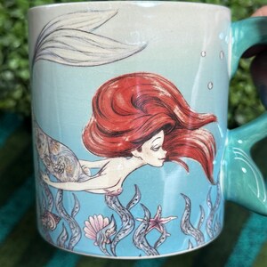 May include: A light blue ceramic mug featuring a graphic illustration of a mermaid with long red hair and a patterned tail. The design includes underwater elements like seaweed, a seashell, and a starfish, with a teal handle shaped like a fin.