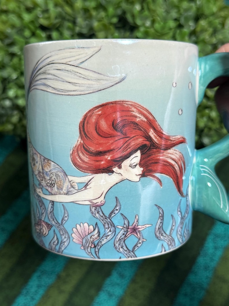 May include: A light blue ceramic mug featuring a graphic illustration of a mermaid with long red hair and a patterned tail. The design includes underwater elements like seaweed, a seashell, and a starfish, with a teal handle shaped like a fin.